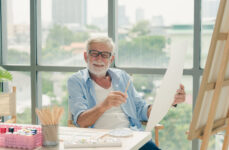 A senior man with glasses holding a small flat brush on his right hand while holding an acrylic paper on his left hand as he smiles directly at the camera