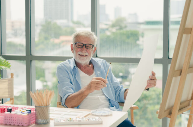 A senior man with glasses holding a small flat brush on his right hand while holding an acrylic paper on his left hand as he smiles directly at the camera