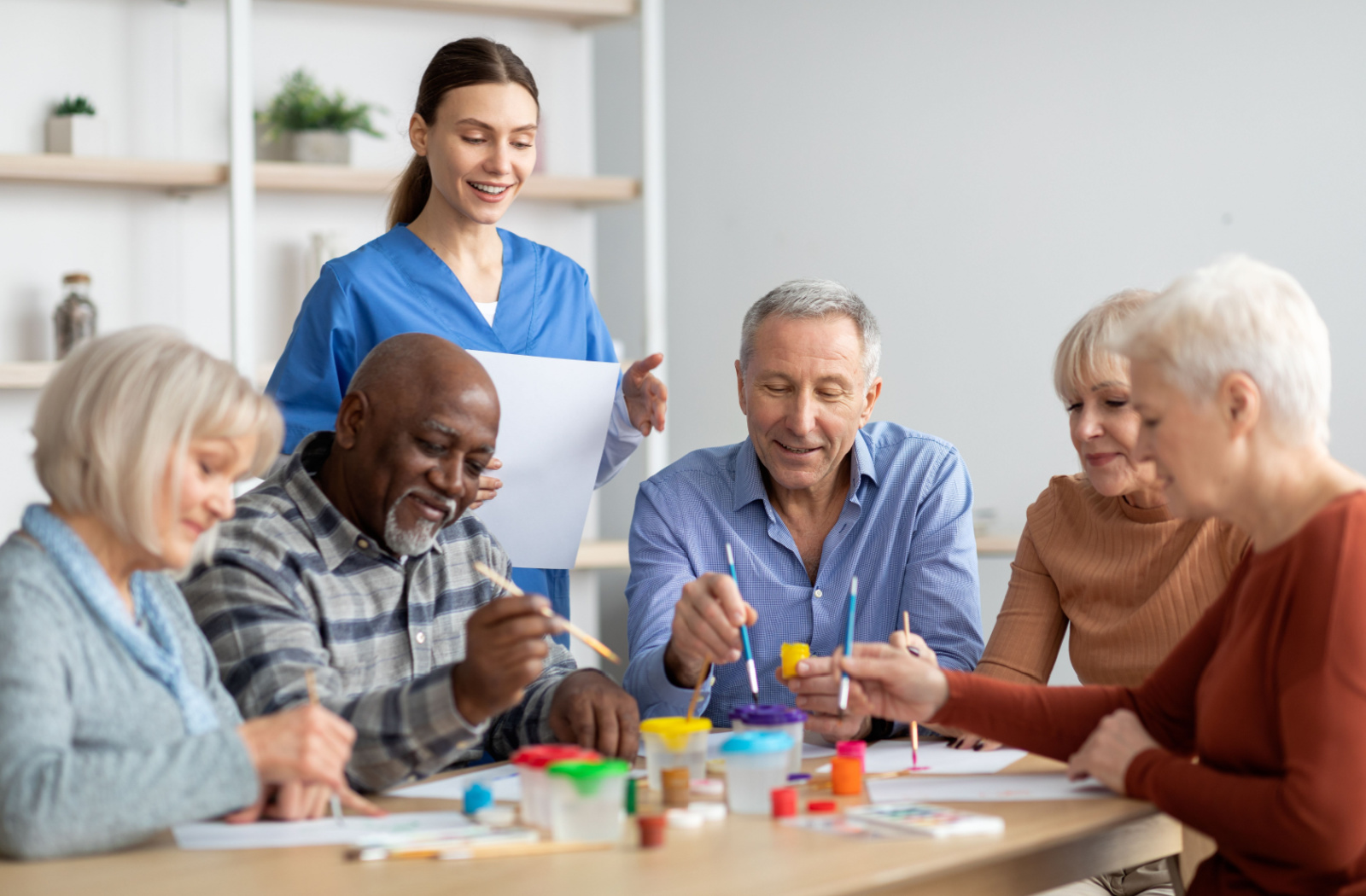 A group of seniors sitting on a table holding a small flat brush and painting, while being supervised by a nurse