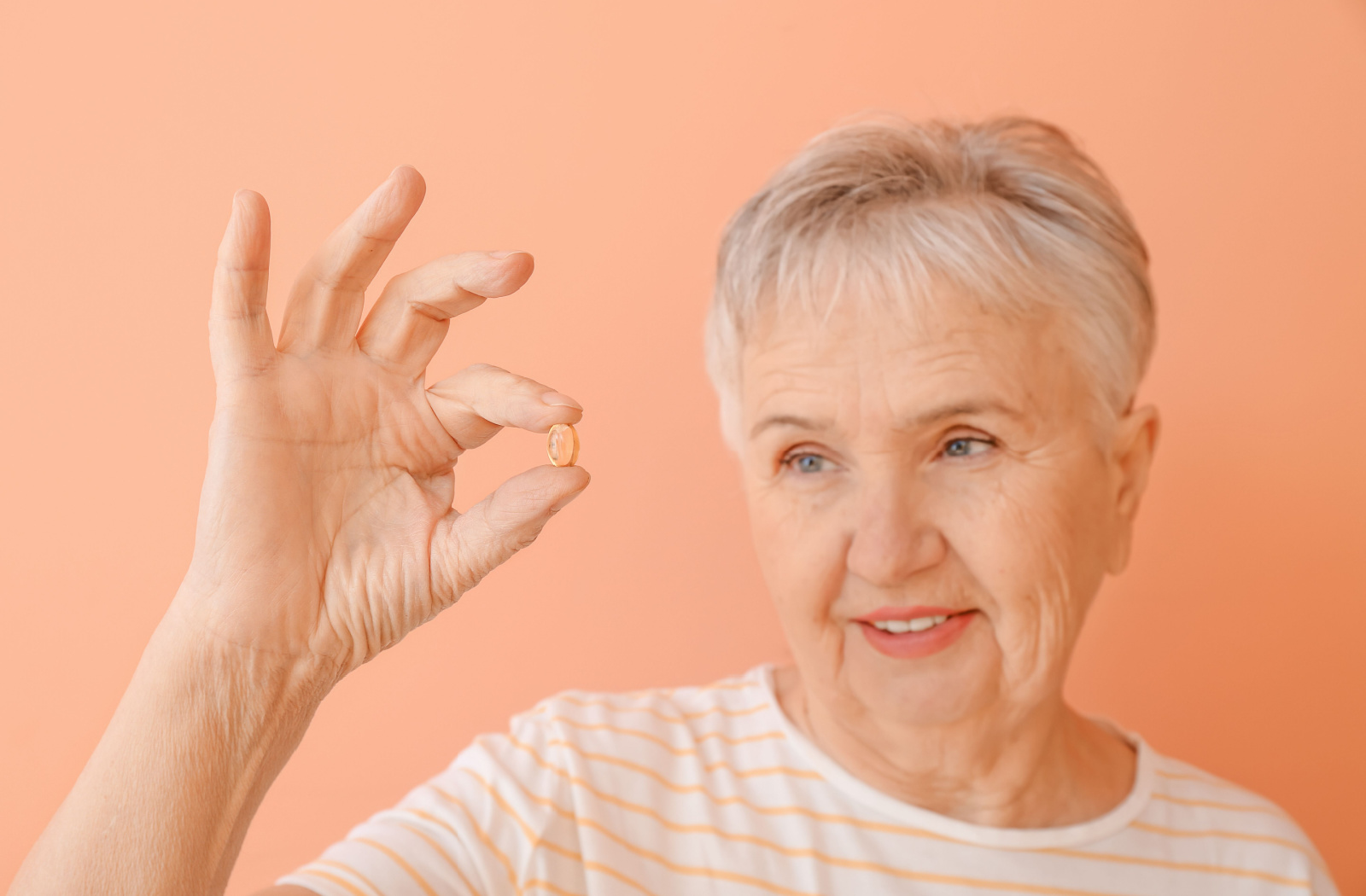 Close-up of a senior woman with short hair holding up a fish oil pill in her right hand against an orange background