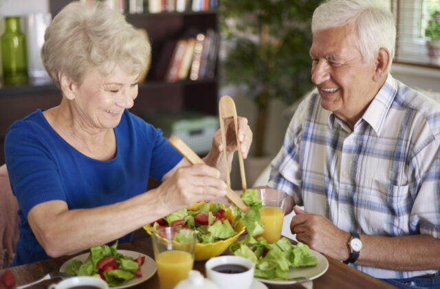A senior woman and man sitting down at a dinner table and smiling while the woman serves salad