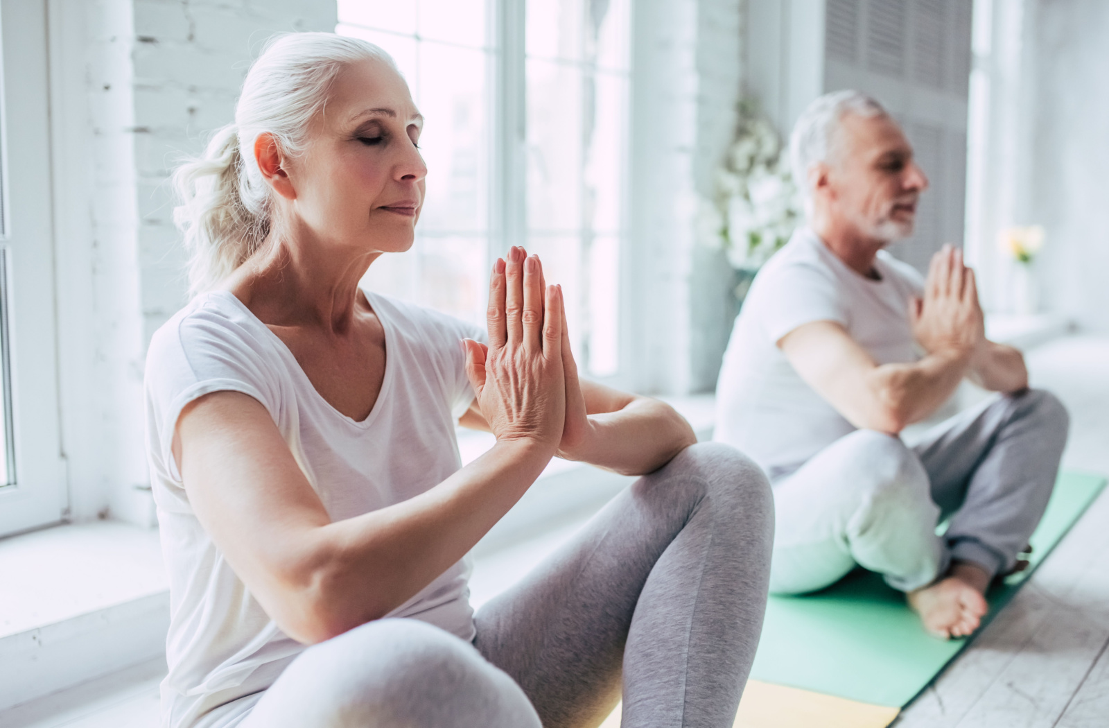 An older adult woman and an older adult man with closed eyes practicing yoga and sitting on yoga mats