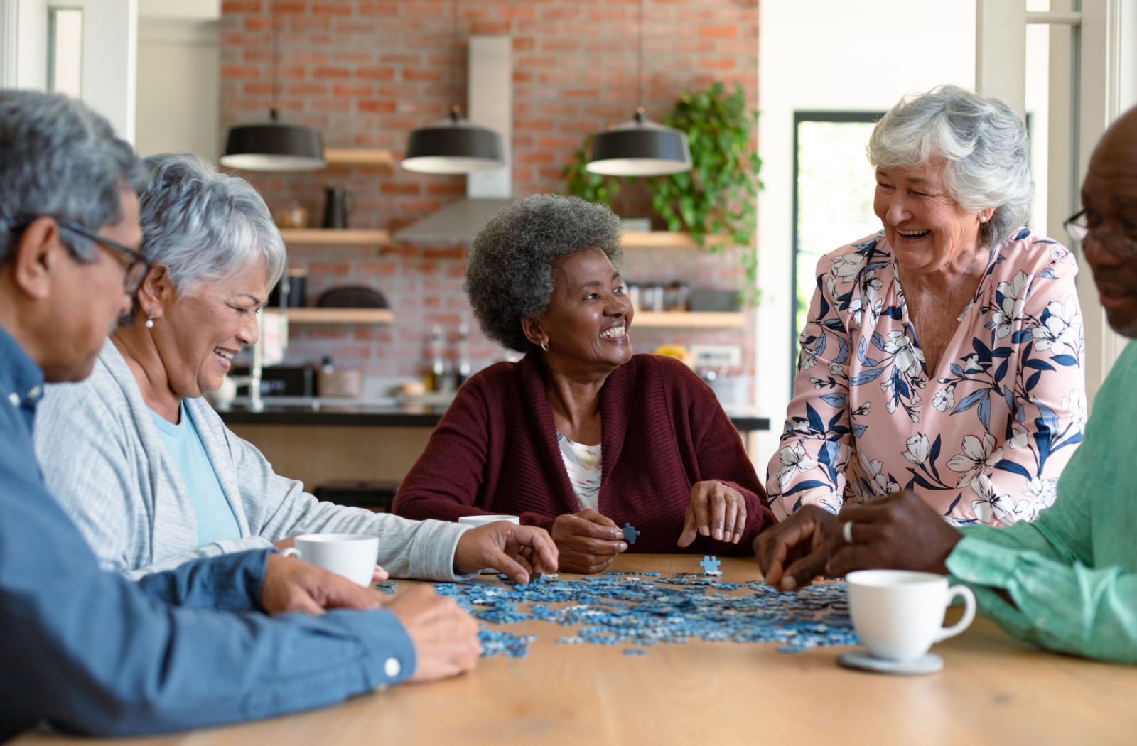 A group of 5 seniors sit at a table drinking coffee while building a puzzle
